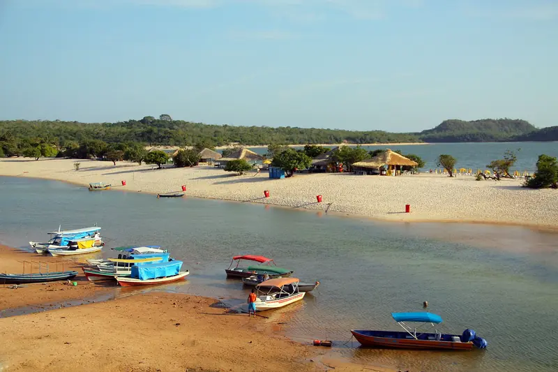 A Ilha do Amor na seca, faixa de areia branca com barracas de palha em frente à vila, barcos coloridos na água rasa e o morro da Serra da Piroca ao fundo