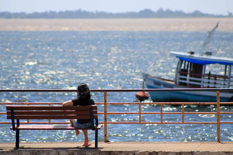 Pessoa sentada num banco na orla, de costas, olhando o rio com uma faixa de água barrenta ao fundo e um barco atracado à direita