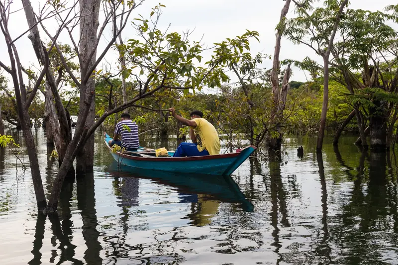 Duas pessoas numa canoa azul entre troncos de árvores dentro da água, numa mata alagada