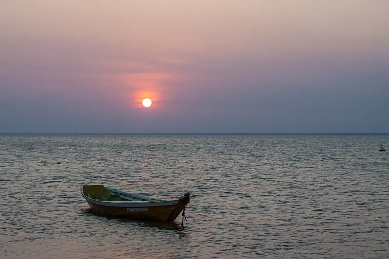 Sol baixo e alaranjado sobre o rio largo e calmo, com uma canoa de madeira ancorada na água rasa