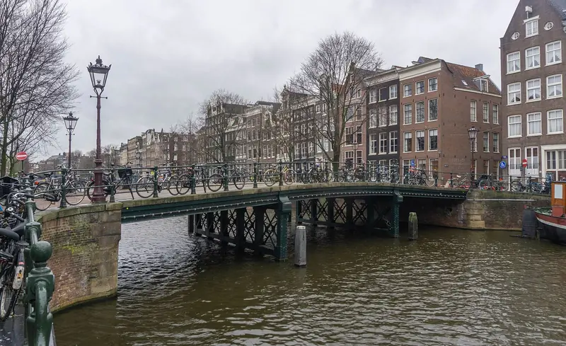 Ponte de ferro verde sobre um canal, com bicicletas presas ao guarda-corpo e casas de tijolo ao fundo num dia nublado
