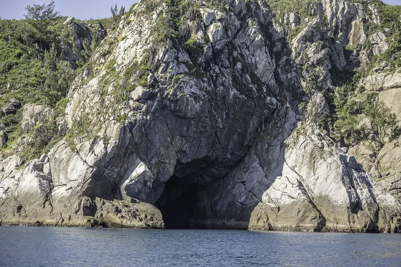Entrada da Gruta Azul, uma abertura escura no costão de pedra clara da Ilha do Cabo Frio, com cactos no alto