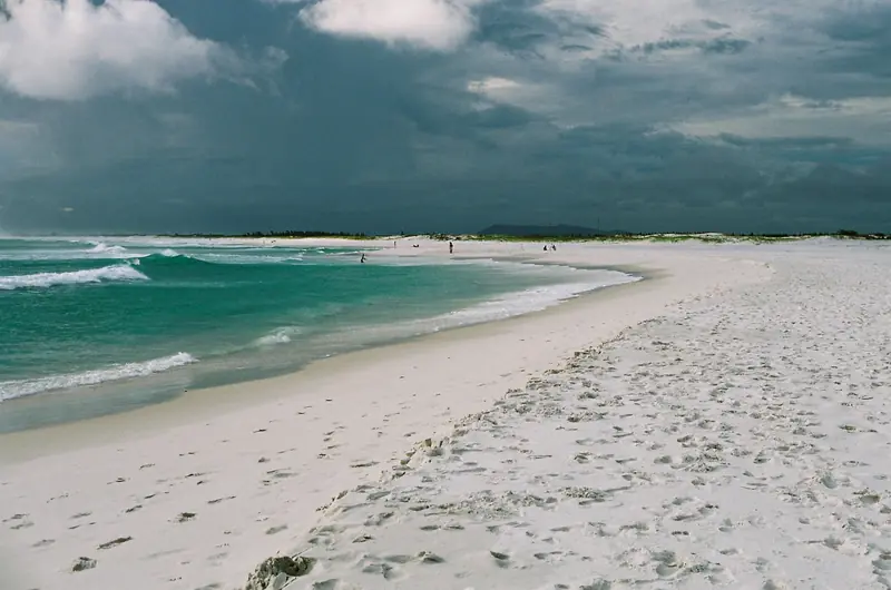 Praia Grande no fim da tarde, com areia branca e larga, marcas de pegadas e ondas verdes quebrando