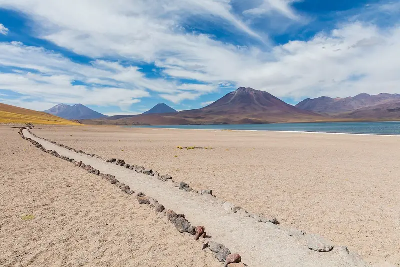Trilha demarcada por pedras atravessando a areia até a Laguna Miscanti, com vulcões ao fundo