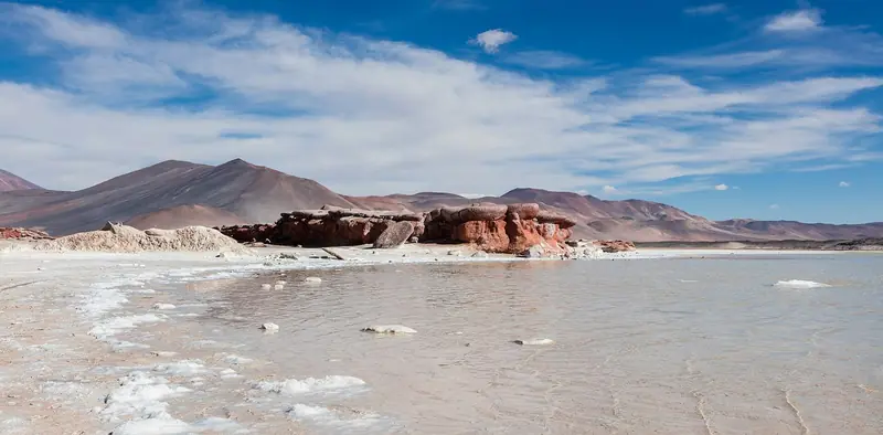 Rochas avermelhadas à beira do Salar de Aguas Calientes Sur, com crostas de sal na água rasa e montanhas ao fundo