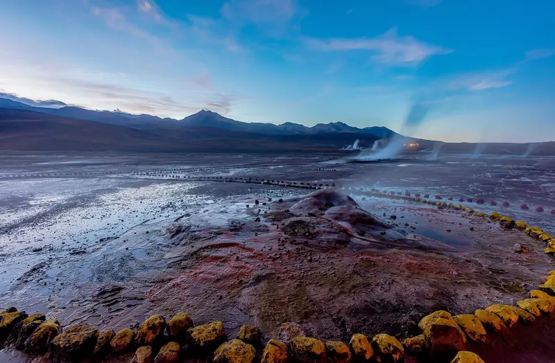 Cone de gêiser com crostas minerais avermelhadas, cercado de pedras, e colunas de vapor ao fundo no amanhecer do Tatio