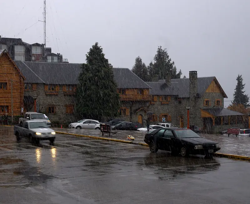 Prédios de pedra e madeira do Centro Cívico de Bariloche sob neve que cai, com carros na praça molhada