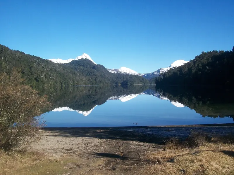 Lago Espejo Chico parado como espelho, refletindo a floresta e três picos nevados sob céu sem nuvens