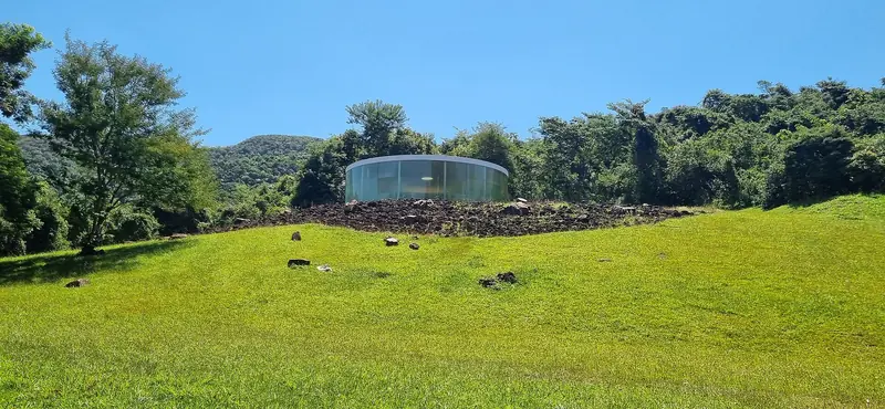 Pavilhão circular de vidro no alto de um gramado, cercado de mata