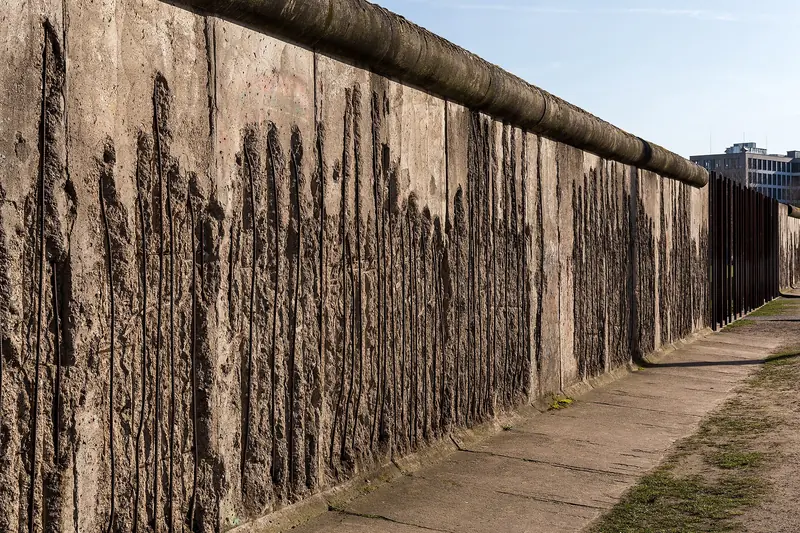 Trecho original do Muro de Berlim, de concreto cinza corroído, ao lado de uma fileira de hastes de ferro que continua a linha da fronteira