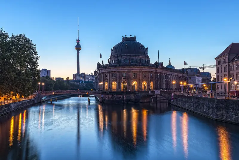 O Bode-Museum, com as janelas iluminadas, na ponta da Ilha dos Museus, refletido no Spree ao amanhecer, com a torre de TV ao fundo