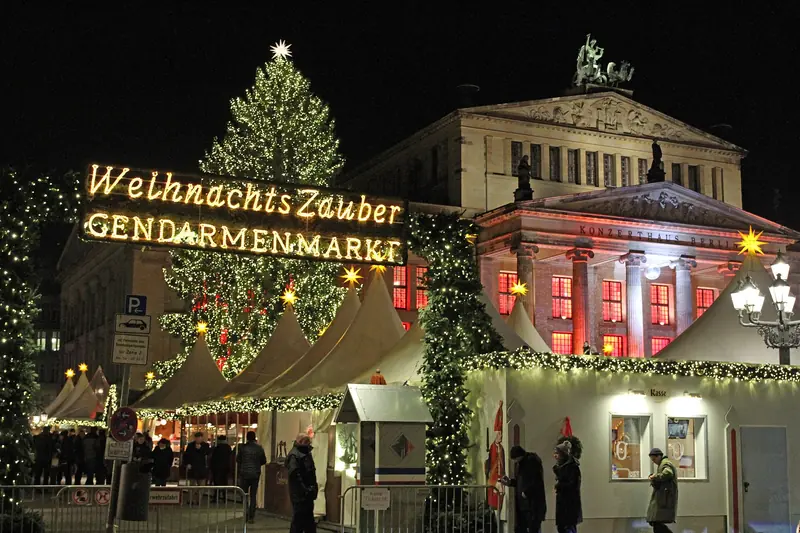 Mercado de Natal do Gendarmenmarkt à noite, com letreiro luminoso, árvore iluminada e barracas brancas diante da Konzerthaus