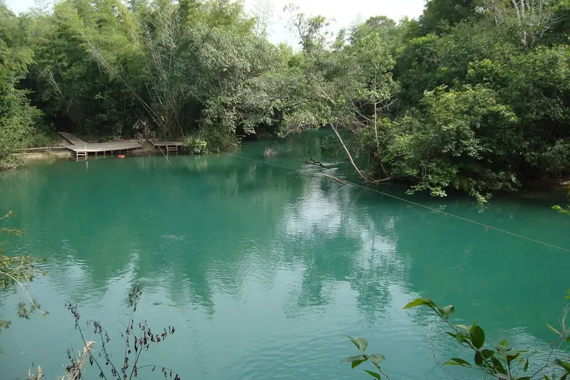Rio de água verde-azulada cercado de mata, com um deque de madeira na margem e um cabo atravessando o rio