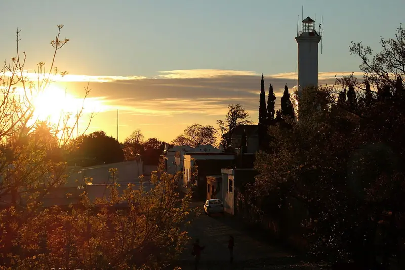 Farol de Colonia del Sacramento contra o céu do fim de tarde, com o sol baixo entre árvores e casas do bairro histórico