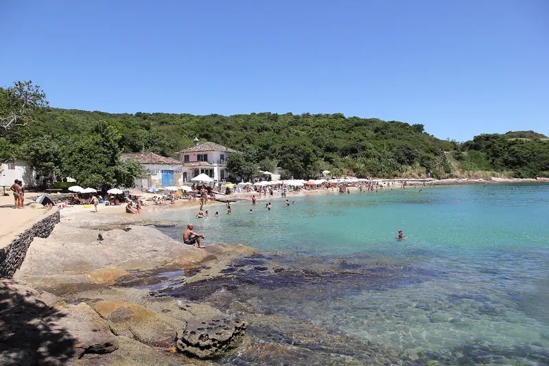 Pedras planas e água verde-clara na Praia da Azeda, com a areia e os guarda-sóis ao fundo