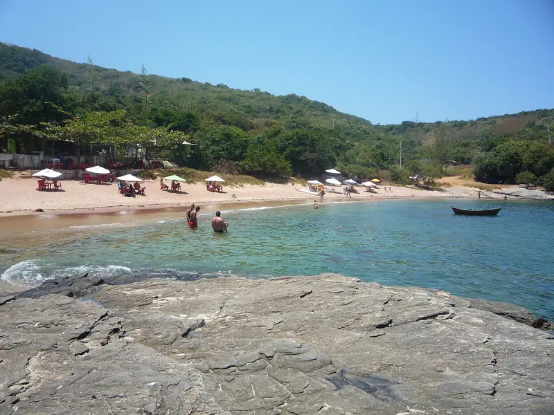 Praia do Forno com areia avermelhada, guarda-sóis e mar calmo entre morros, vista das pedras do canto