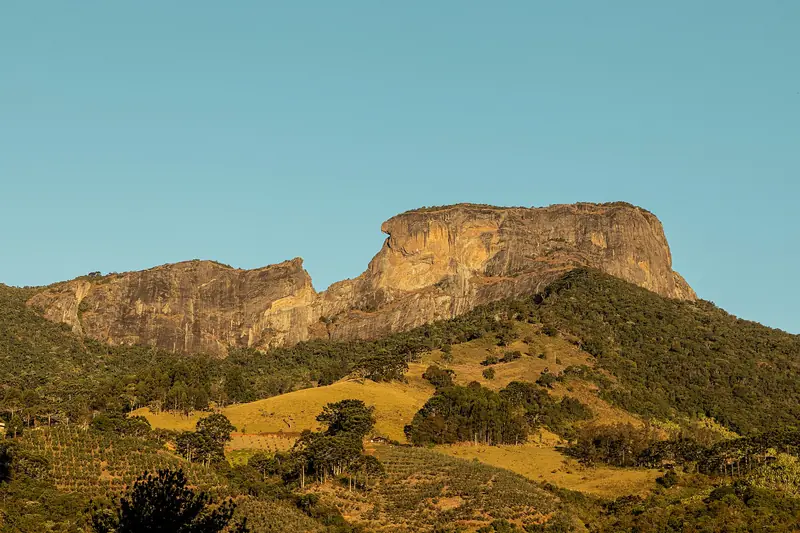 Paredão de rocha da Pedra do Baú sobre morros com mata e pasto, sob céu azul