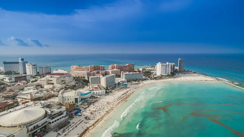 Vista aérea de Punta Cancún, com hotéis na ponta da Zona Hotelera, praia de areia branca em curva e mar turquesa com faixas escuras de sargaço