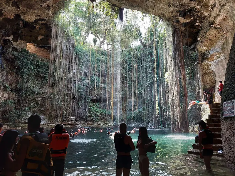 Cenote Ik Kil visto de dentro, com raízes e cipós descendo das bordas até a água e banhistas de colete na parte rasa