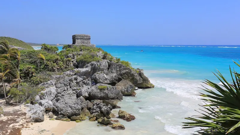 Templo do Deus do Vento em Tulum sobre um promontório de pedra, com palmeiras e mar azul-turquesa ao fundo