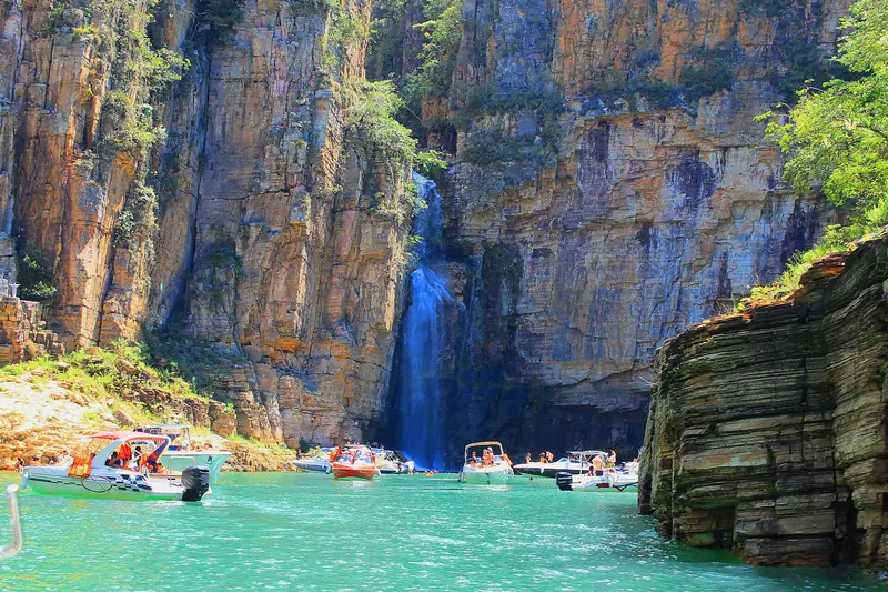 Lanchas paradas ao pé de uma cachoeira fina que desce de um paredão alaranjado, com água verde-turquesa em primeiro plano