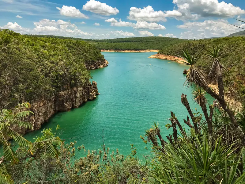 Braço de lago verde-turquesa entre margens de mato, com uma faixa de areia clara exposta na beira da água