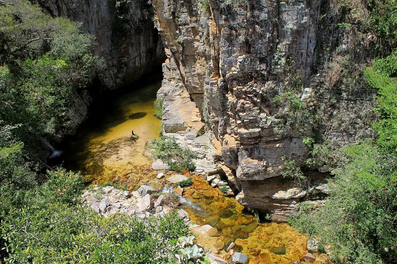 Vista do alto de um cânion estreito de rocha cinza, com um poço de fundo dourado e pedras claras na saída