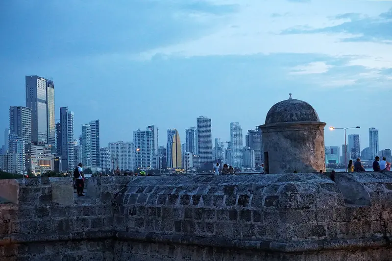 Torre de vigia de pedra sobre a muralha de Cartagena ao anoitecer, com os prédios altos de Bocagrande ao fundo e pessoas sentadas no parapeito