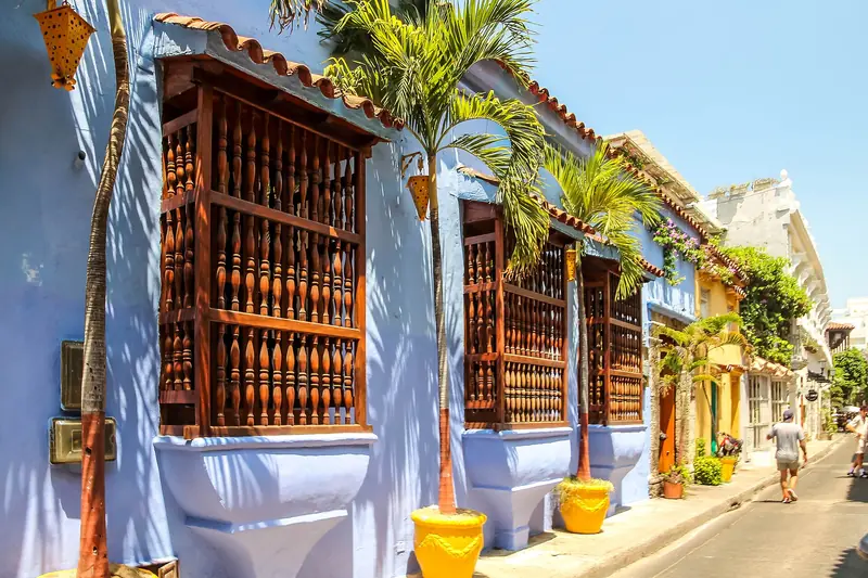 Rua da Cidade Murada de Cartagena com casas pintadas de azul, janelas salientes de balaústres de madeira, palmeiras em vasos amarelos e um pedestre ao fundo