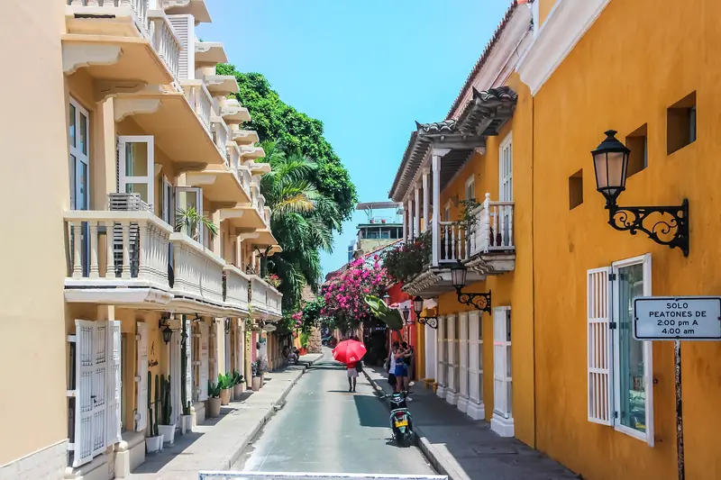 Rua da Cidade Murada com sobrados de sacadas brancas à esquerda, casa amarela com lampião de ferro à direita, buganvílias rosa ao fundo e placa de rua exclusiva para pedestres