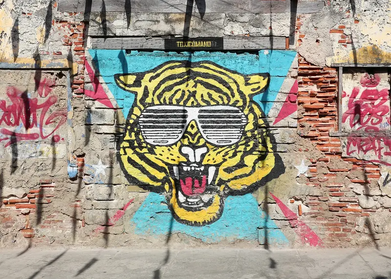 Mural de um tigre amarelo de óculos escuros pintado numa parede de tijolos descascados, cercado de pichações