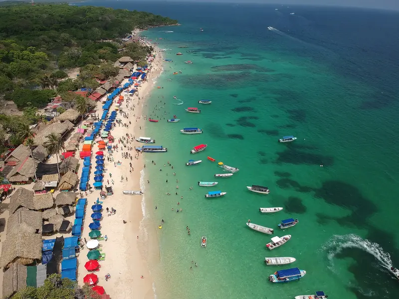 Vista aérea de Playa Blanca com faixa de areia branca tomada por guarda-sóis e cabanas, mar verde-claro e dezenas de lanchas ancoradas perto da praia