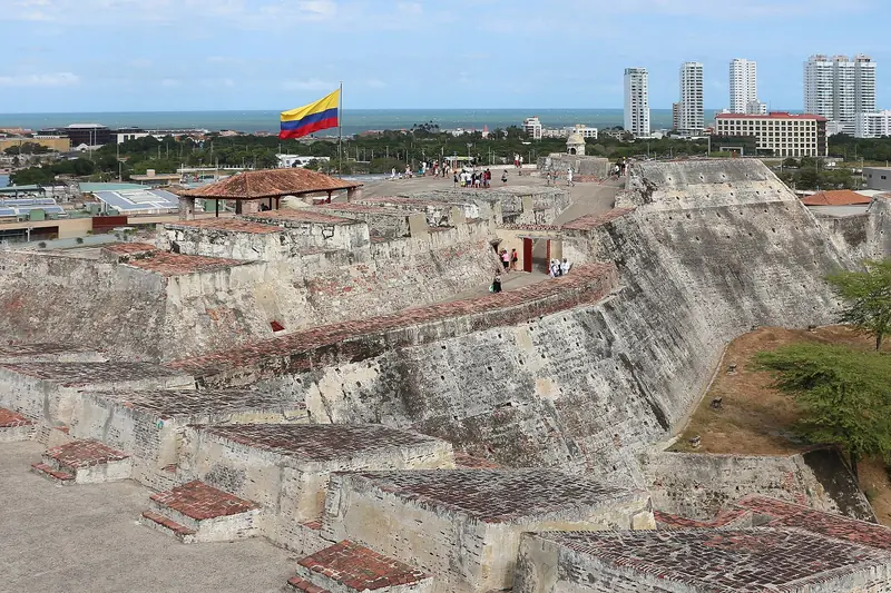 Rampas e muralhas de pedra do Castillo de San Felipe vistas do alto, com bandeira da Colômbia, visitantes no topo e prédios da cidade ao fundo