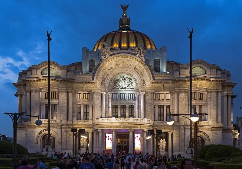 Fachada de mármore do Palácio de Bellas Artes iluminada ao anoitecer, com a cúpula dourada sob céu azul-escuro e pessoas na praça em frente