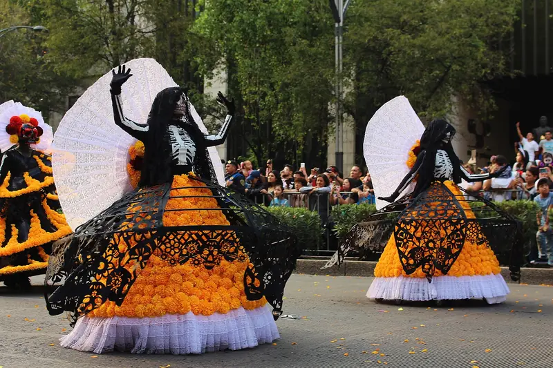 Catrinas com rosto pintado de caveira, leques brancos e saias armadas de flores de cempasúchil sob renda preta, desfilando na Reforma diante do público