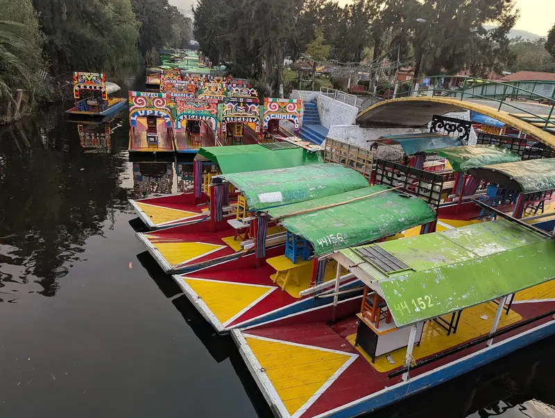 Trajineras coloridas atracadas em fila num embarcadouro de Xochimilco, com toldos verdes e cascos pintados de vermelho e amarelo