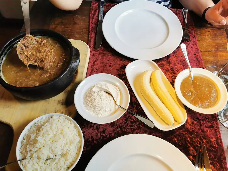 Panela de barro com carne desfiada ao lado de farinha de mandioca, bananas e arroz
