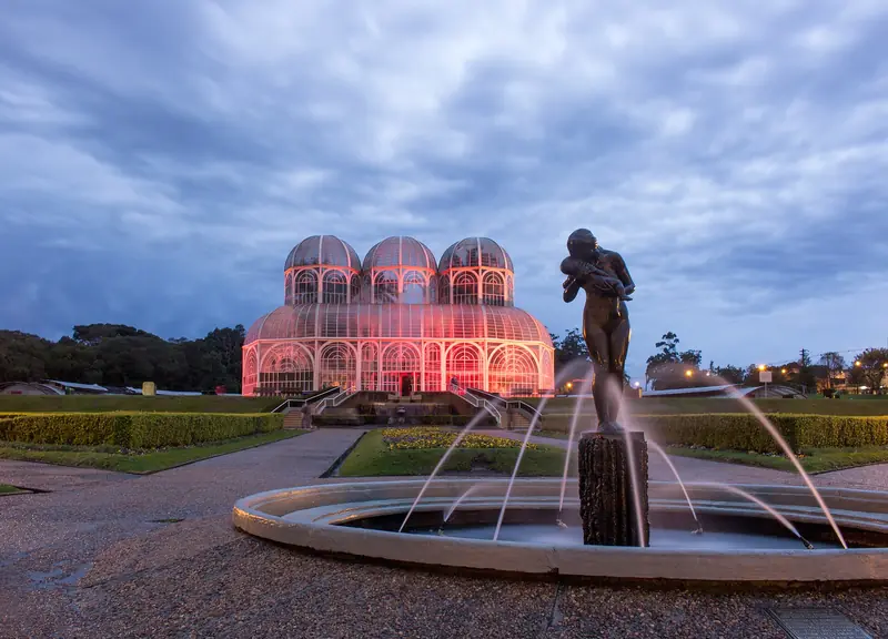 Estufa de ferro e vidro do Jardim Botânico de Curitiba iluminada ao anoitecer, com uma fonte e uma estátua em primeiro plano