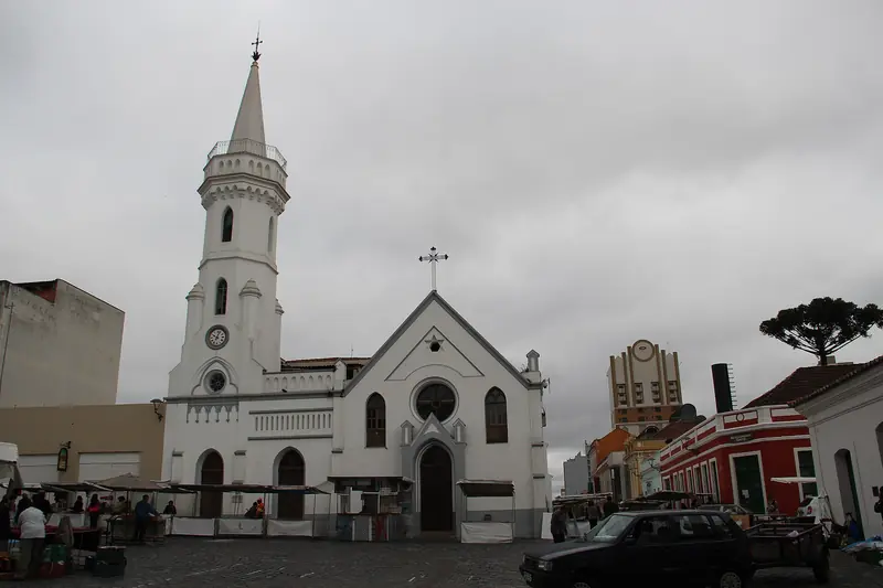 Igreja branca com torre e relógio no Largo da Ordem, com barracas da feira montadas na praça de pedra