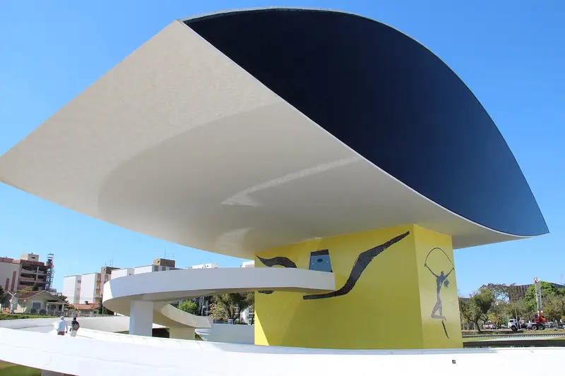 Prédio do Museu Oscar Niemeyer em forma de olho, sobre um pilar amarelo, com rampa branca e céu azul