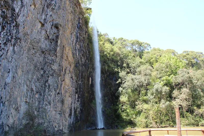 Cascata caindo do paredão de rocha de uma antiga pedreira, com mata ao lado e um lago embaixo