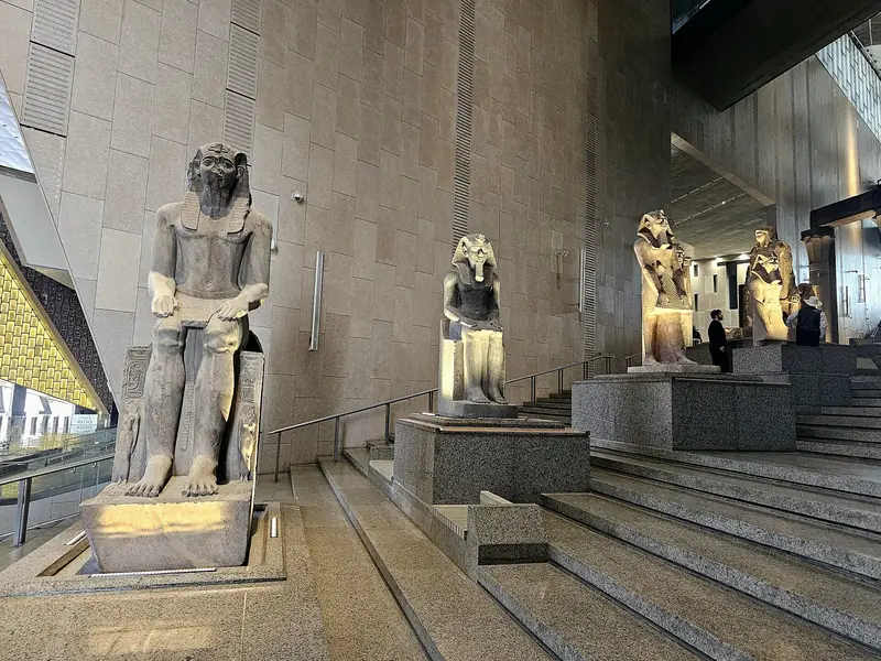 Estátuas de faraós sentados e de pé em pedestais ao longo dos degraus largos de uma escadaria interna, com paredes altas de pedra clara