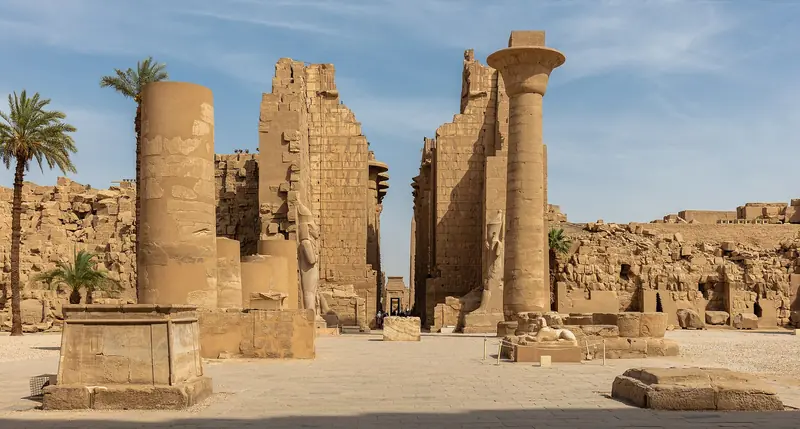 Pátio de Karnak com uma coluna isolada à direita, um portal de pedra em ruínas ao centro e palmeiras à esquerda, sob céu azul