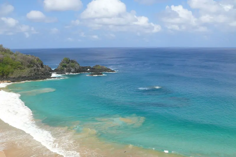 Baía do Sancho com água verde e azul, faixa de areia clara e a costa recortada ao fundo