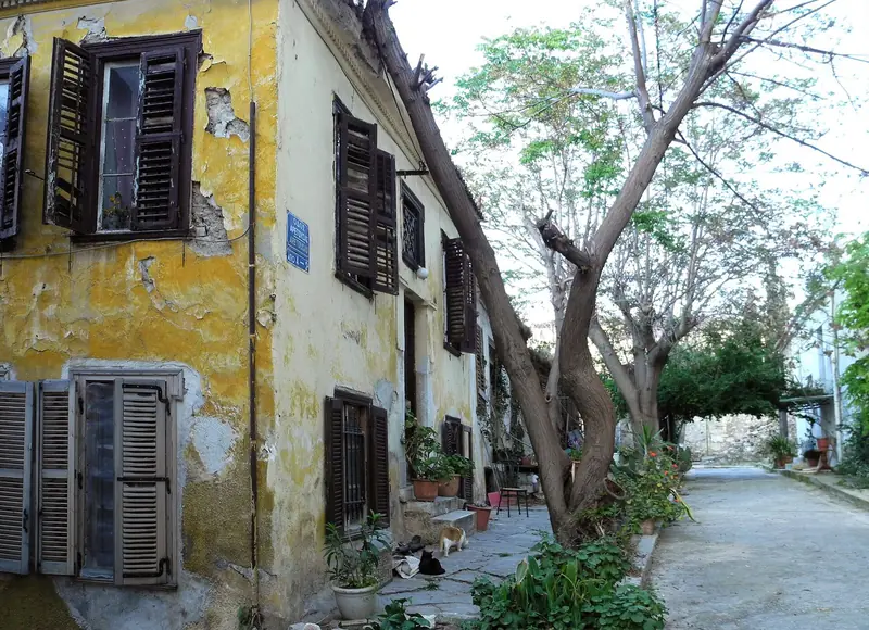 Casa amarela de venezianas de madeira na rua Aretousas, em Anafiotika, com vasos de plantas e dois gatos na calçada
