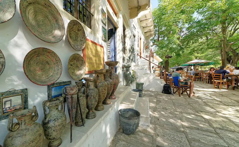 Calçada de mármore em Apiranthos, com cerâmicas penduradas na parede branca e mesas de taverna à sombra das árvores