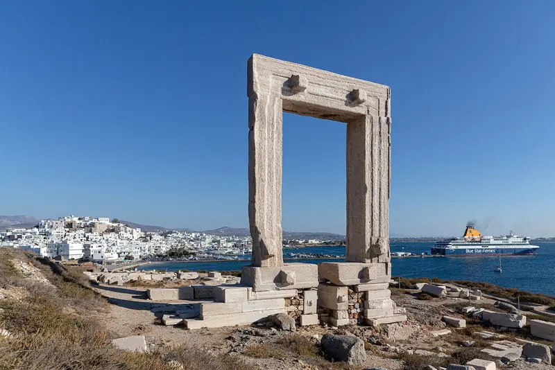 A Portara, portal de mármore numa ilhota, com a Chora de Naxos ao fundo e um ferry da Blue Star chegando ao porto