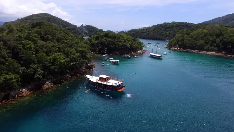 Vista aérea da Lagoa Azul, canal de água verde-azulada entre a costa da ilha e ilhotas de mata, com escunas ancoradas e gente nadando