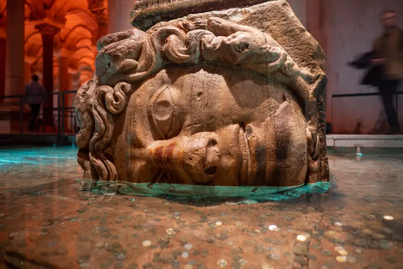 Cabeça de Medusa de mármore deitada de lado na base de uma coluna, com moedas no chão alagado da Cisterna da Basílica