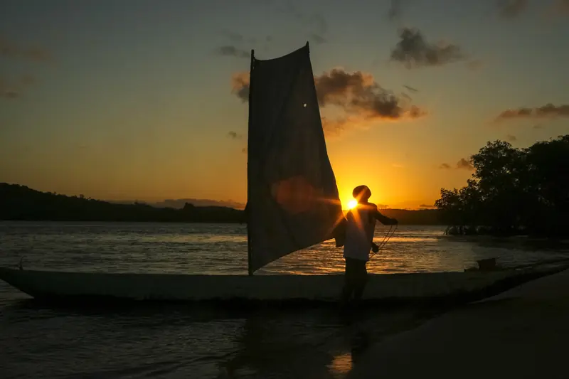 Silhueta de um homem ao lado de uma canoa com vela, na beira do rio, com o sol se pondo atrás da mata na outra margem